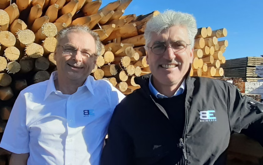 Deux hommes souriants devant des troncs d'arbres.